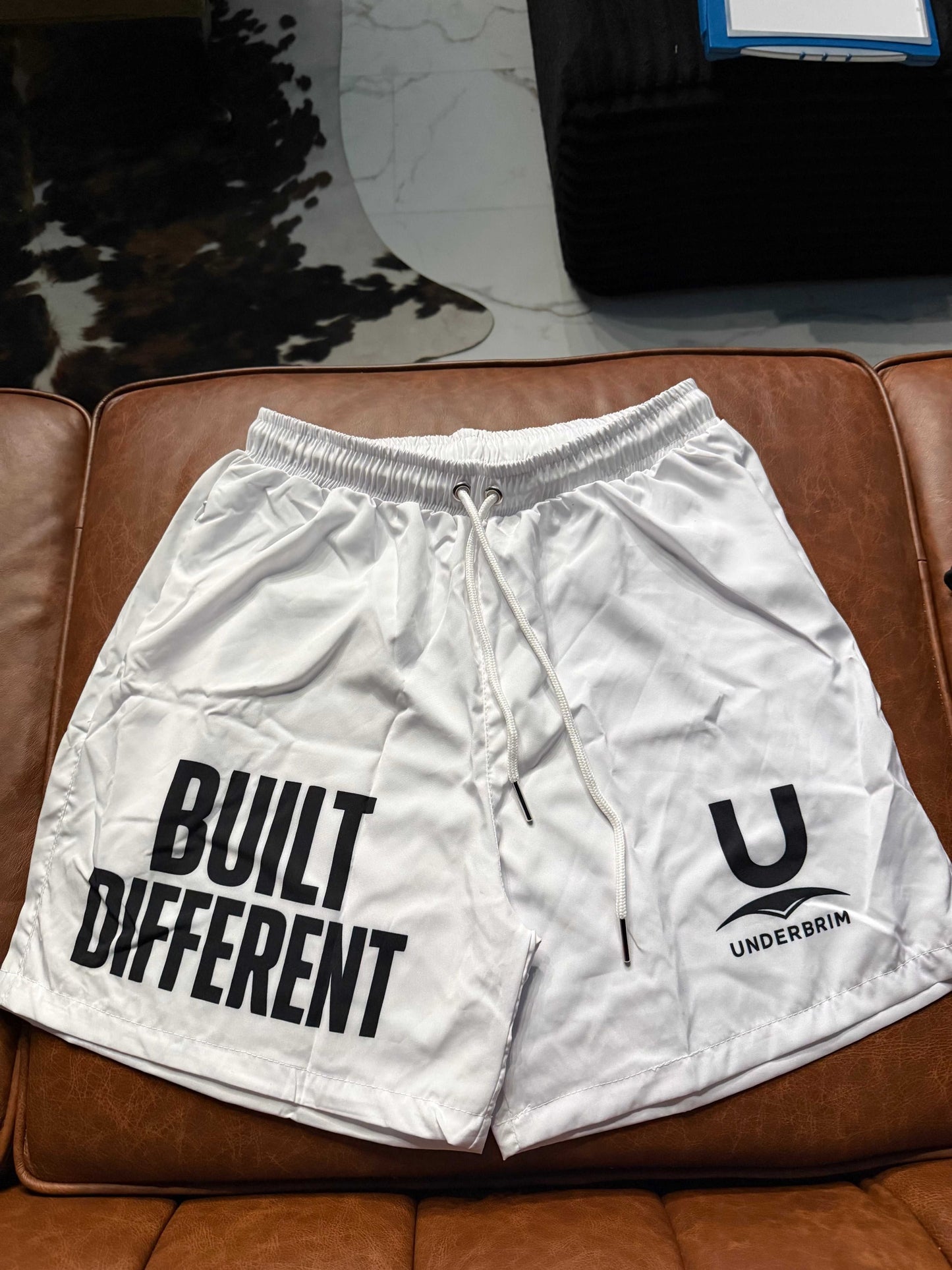 White Underbrim Built Different shorts with bold 'BUILT DIFFERENT' text and U logo, drawstring waistband.